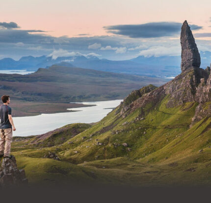 A person enjoying a scenic view of lush hills and a loch at sunset near a towering rock formation.