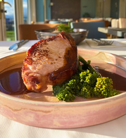 A delicious pork dish with sauce and broccoli in a sunlit Balmacara hotel dining area.