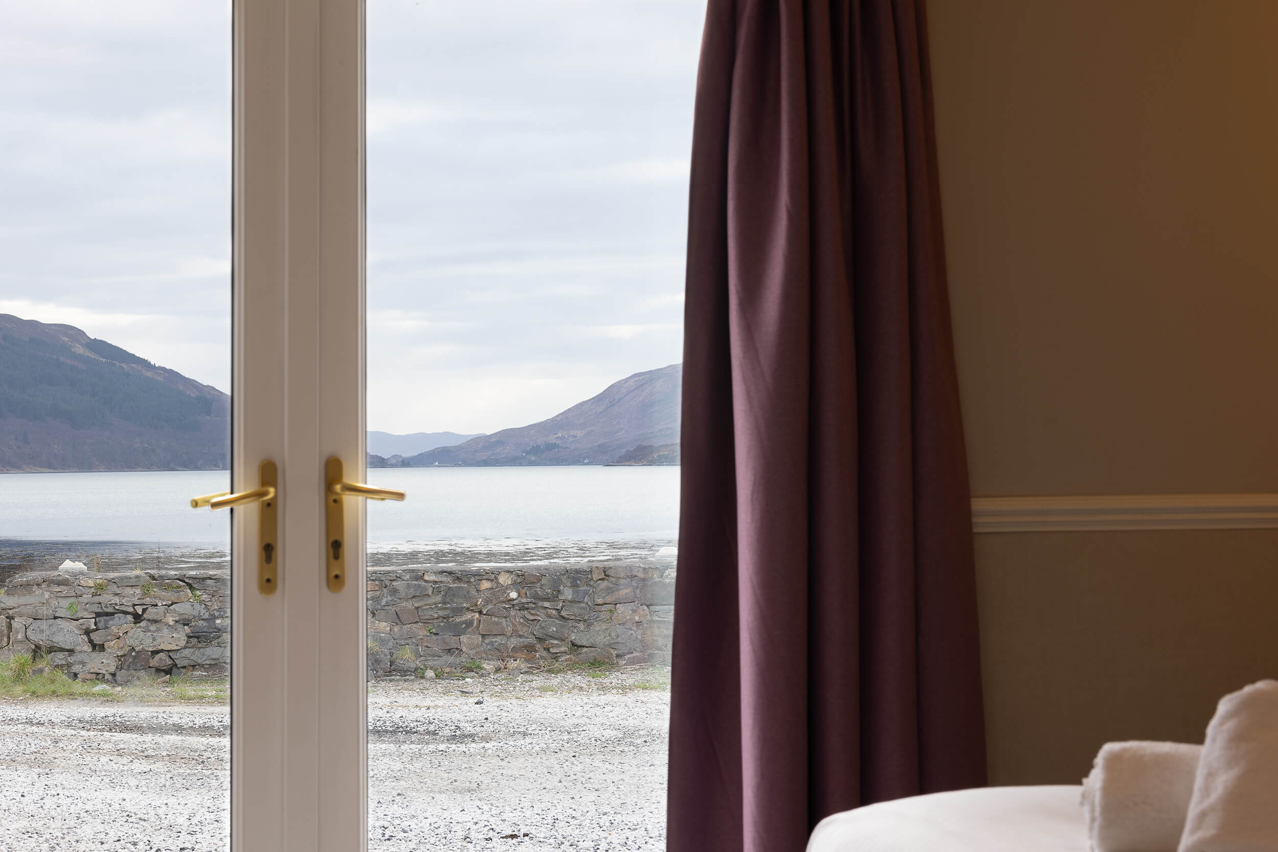 View of serene loch and mountains from hotel room, framed by elegant purple curtains.