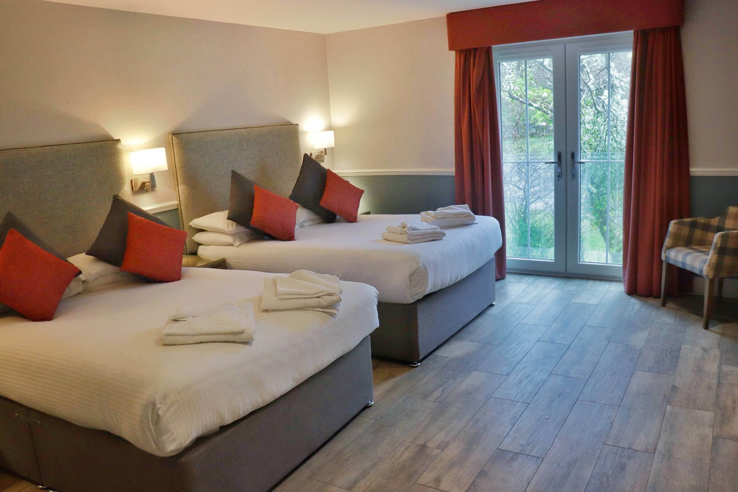 Spacious hotel room with two cosy double beds, red and grey pillows, and garden view.