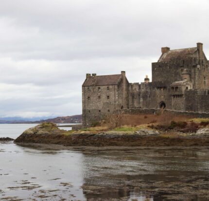 Historic stone castle by a serene loch under cloudy skies, surrounded by mountains.