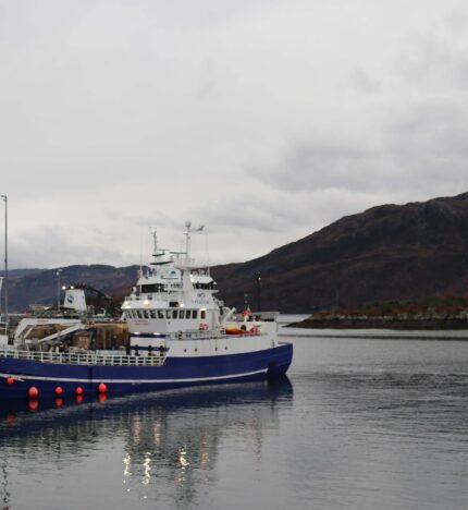 A blue and white boat docked at Balmacara harbour, surrounded by scenic hills under a cloudy sky.