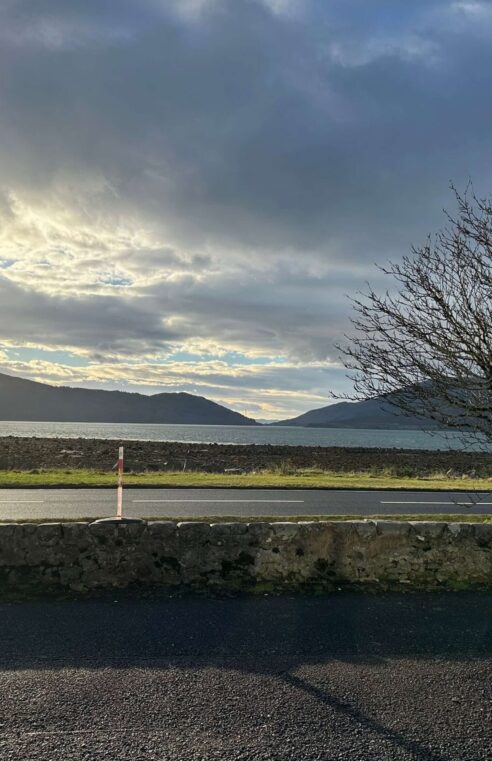 View of a serene loch with a bare tree, mountains, and sunlit clouds in Balmacara.