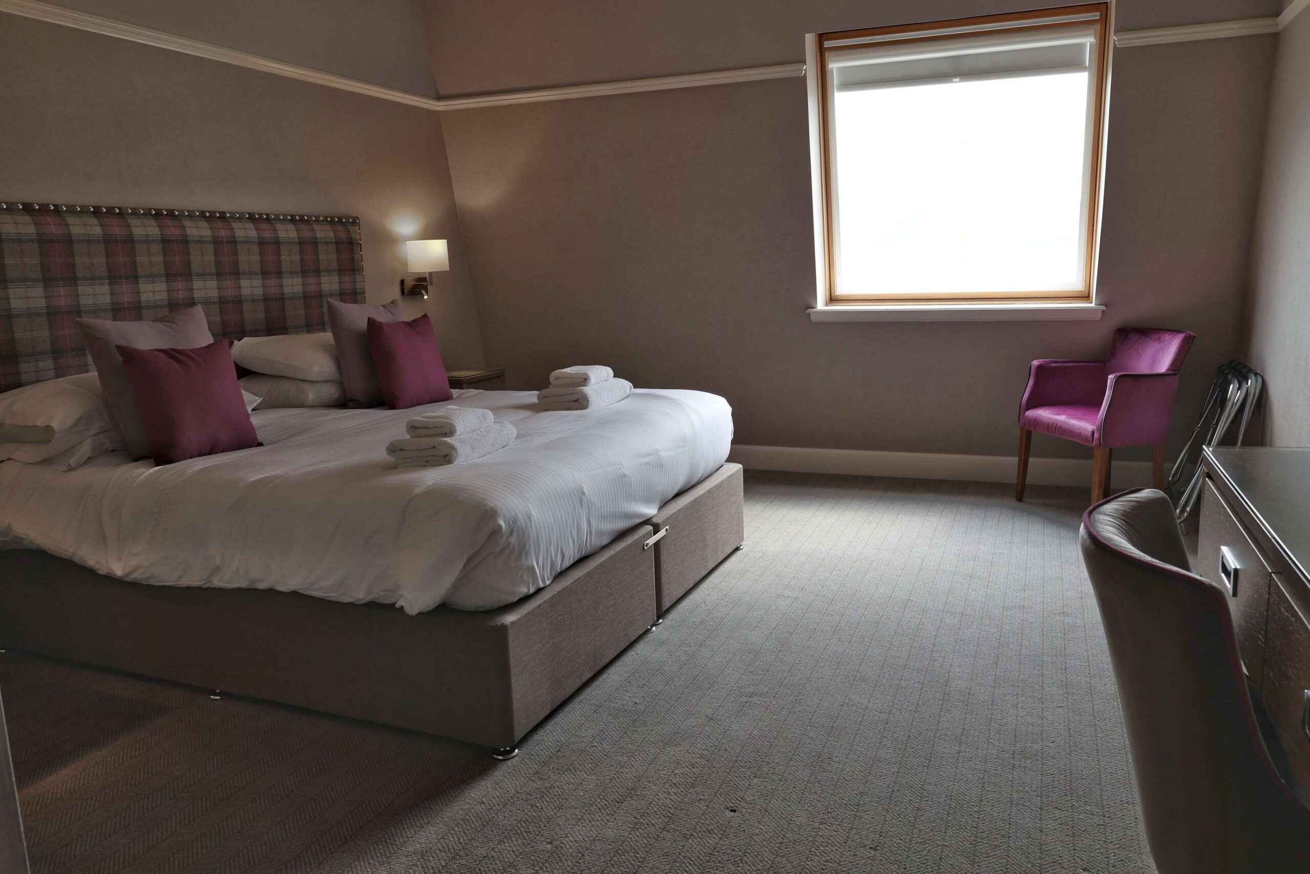 Comfortable hotel room with double bed, purple accents, cosy chair, and soft lighting.